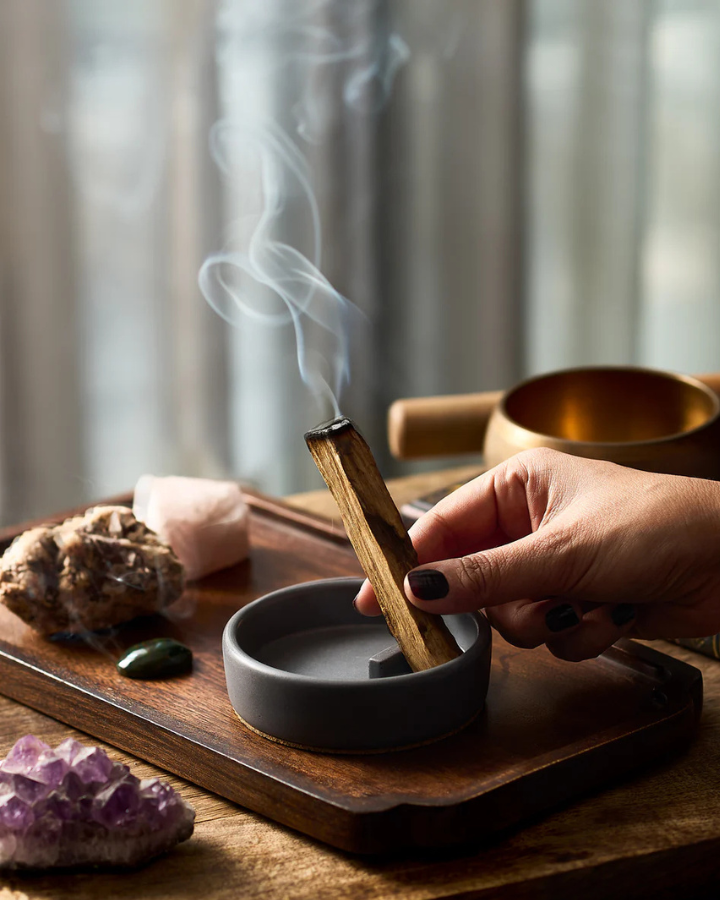 Person holding a Palo Santo over a small bowl with smoke rising, on a wooden tray with crystals.